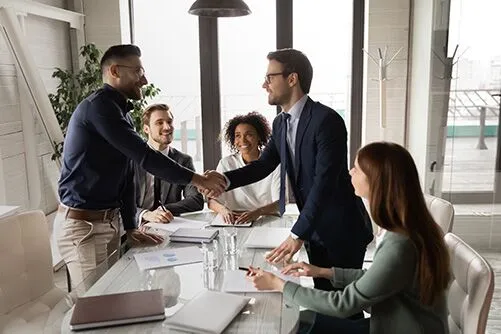 business-meeting-citizenship-by-investment-discussion Two businesspeople shaking hands in a meeting with colleagues discussing citizenship by investment options in a modern office setting.