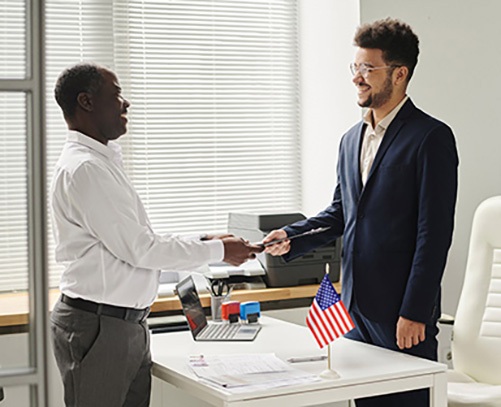 Two professionals shaking hands in an office, symbolizing a successful EB-5 citizenship by investment agreement, with an American flag on the desk.